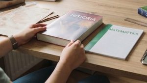 Na obrazie widać jasne, drewniane biurko w pokoju, przy którym siedzi osoba