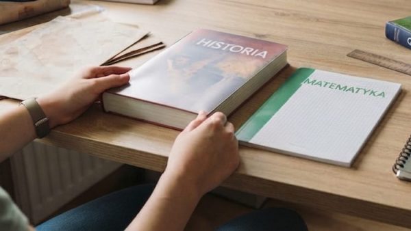 Na obrazie widać jasne, drewniane biurko w pokoju, przy którym siedzi osoba